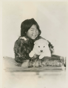 Image of Shoo-e-ging-wa [Suakannguaq Qaerngaaq] and white puppy. Coat a bluish gray. Boots ivory. Suakanquak Kaerngar